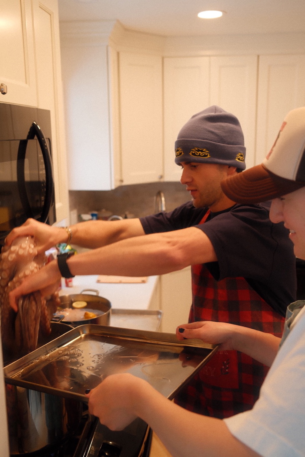 Matt in a beanie and dark tee working alongside another cook, hands on raw octopus over a stainless-steel tray in a home kitchen.